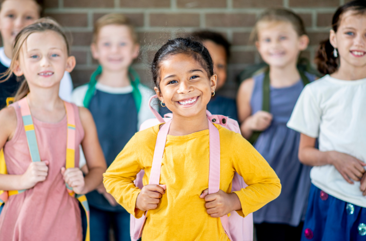 A photo of a little girl wearing a yellow top. Smiling looking directly at the camera. There are other kids in the background the photo is focused on the young girl in the center.