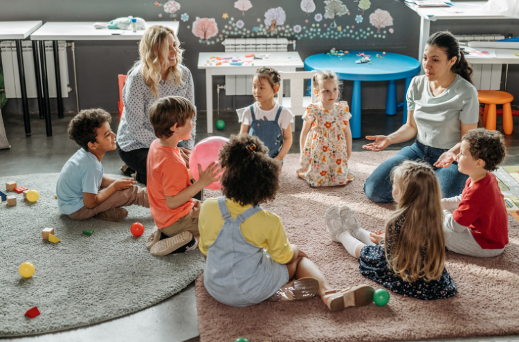A diverse group of young children sitting in a circle on the floor of a bright classroom, engaging in a group activity with two female teachers.