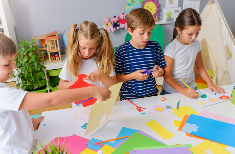 Four young children sitting around a white table filled with colorful construction paper, scissors, and pencils, working on creative arts and crafts projects.